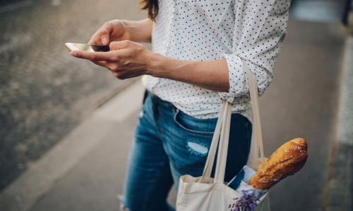 woman walks down the street and texts with a bag of groceries on her arm